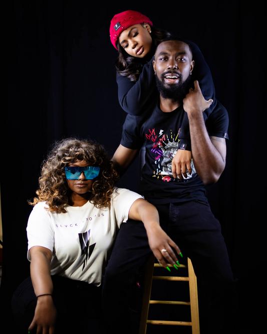 male with 2 woman wearing Black short-sleeve t-shirt featuring a bold, graphic design of a lion king surrounded by symbols of power, knowledge, and sacrifice. The text "Only the Hungry Eat with Kings" is prominently displayed, along with the Blvck Exodus logo.girl on male shoulder has red distressed beanie and woman on right have white graphic T shirt