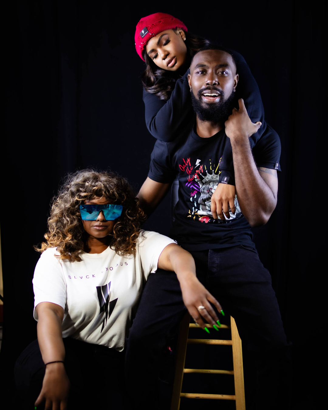 male with 2 woman wearing Black short-sleeve t-shirt featuring a bold, graphic design of a lion king surrounded by symbols of power, knowledge, and sacrifice. The text "Only the Hungry Eat with Kings" is prominently displayed, along with the Blvck Exodus logo.girl on male shoulder has red distressed beanie and woman on right have white graphic T shirt 